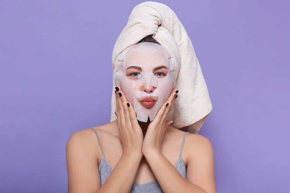 Woman wearing face sheet mask with towel on head in front of purple background