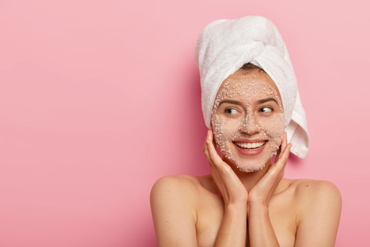 Woman with exfoliant on face wearing towel in front of pink backdrop