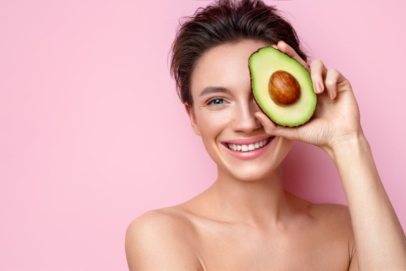 Woman smiling and holding half an avocado over eye in front of pink backdrop