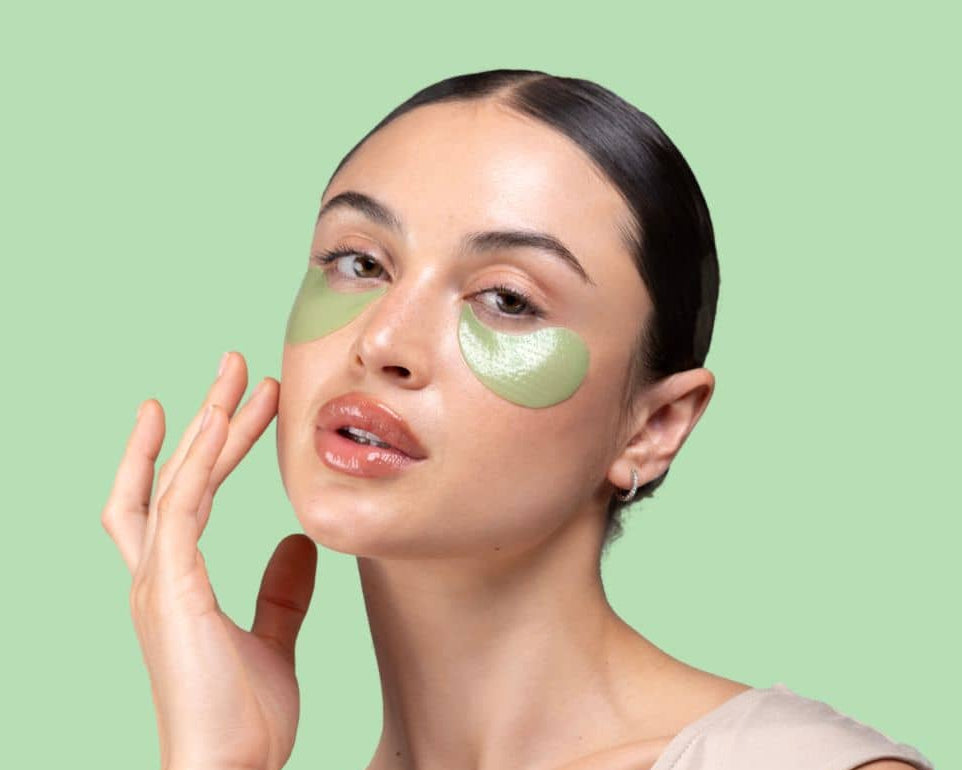 Woman applying green cool crush eye patches on a light green background