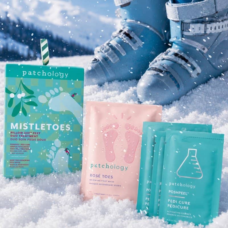 Foot care masks and exfoliating peel displayed in the snow: a teal 'Mistletoes' foot soak box, a pink Several teal single-use packets, with blue ski boots in the background.