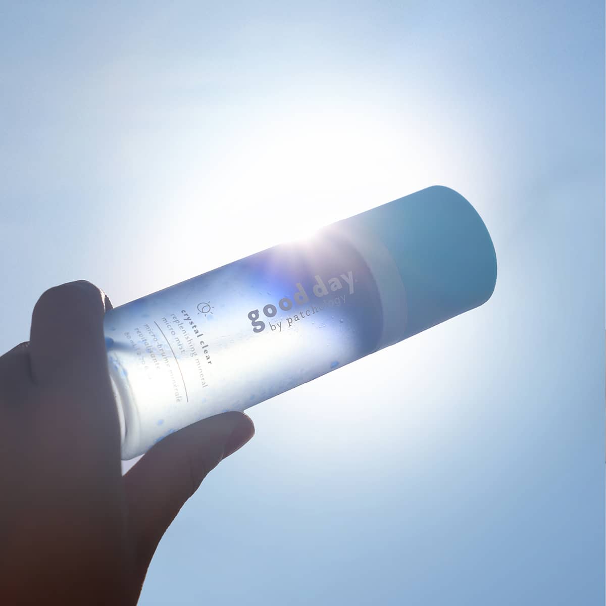 A hand holds a cylindrical bottle of good day crystal clear mist against a bright sunlit sky, with strong backlighting and lens flare.