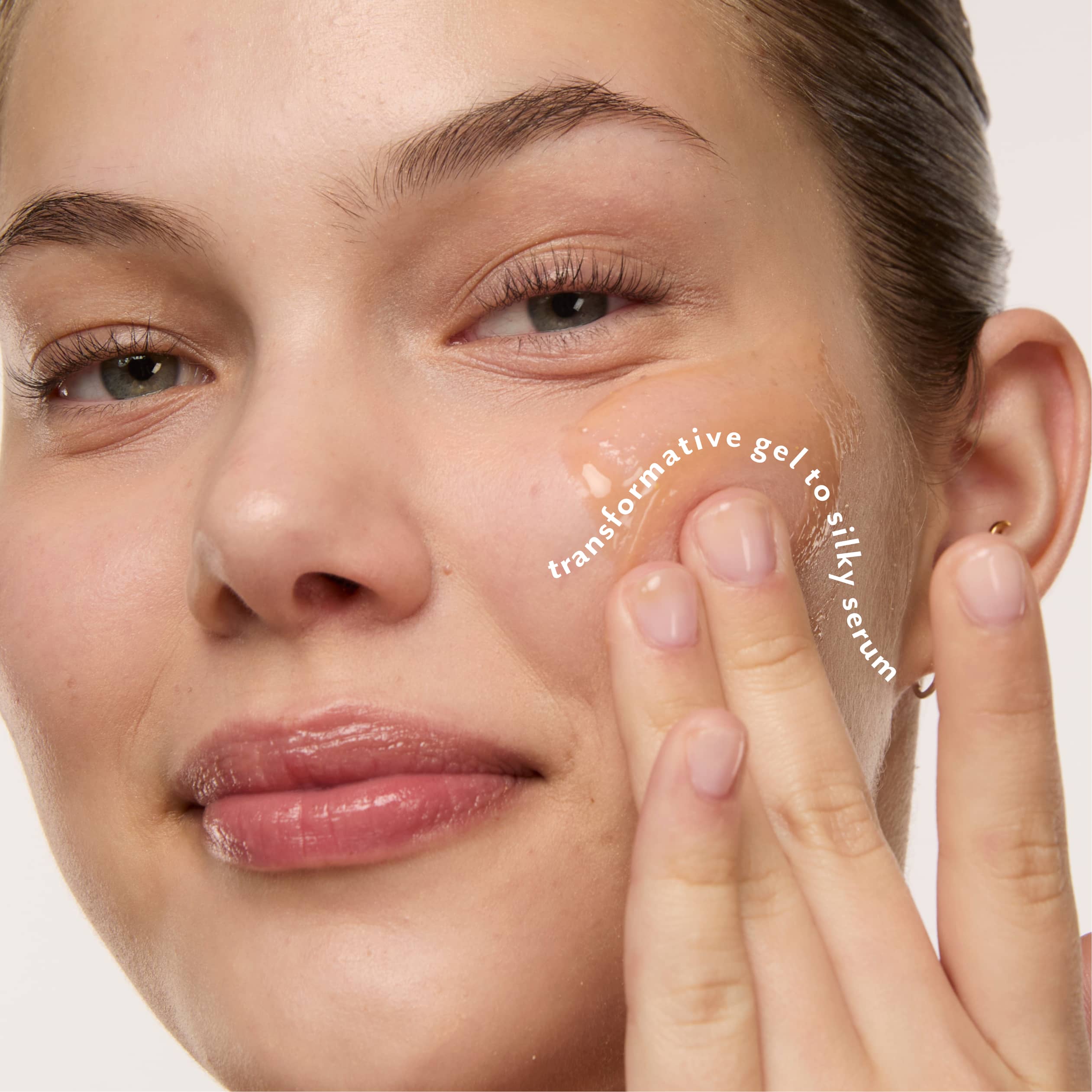 Close-up of a woman applying a glossy, gel serum to her cheek with her finger. White text on the cheek reads 'transformative gel to silky serum'.