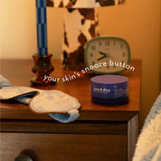 A small round cosmetic jar labeled 'good day' sits on a wooden bedside table next to a blue-and-white eye mask, with a lamp, candle, and clock in the background. White text across the image reads 'your skin's snooze button'.