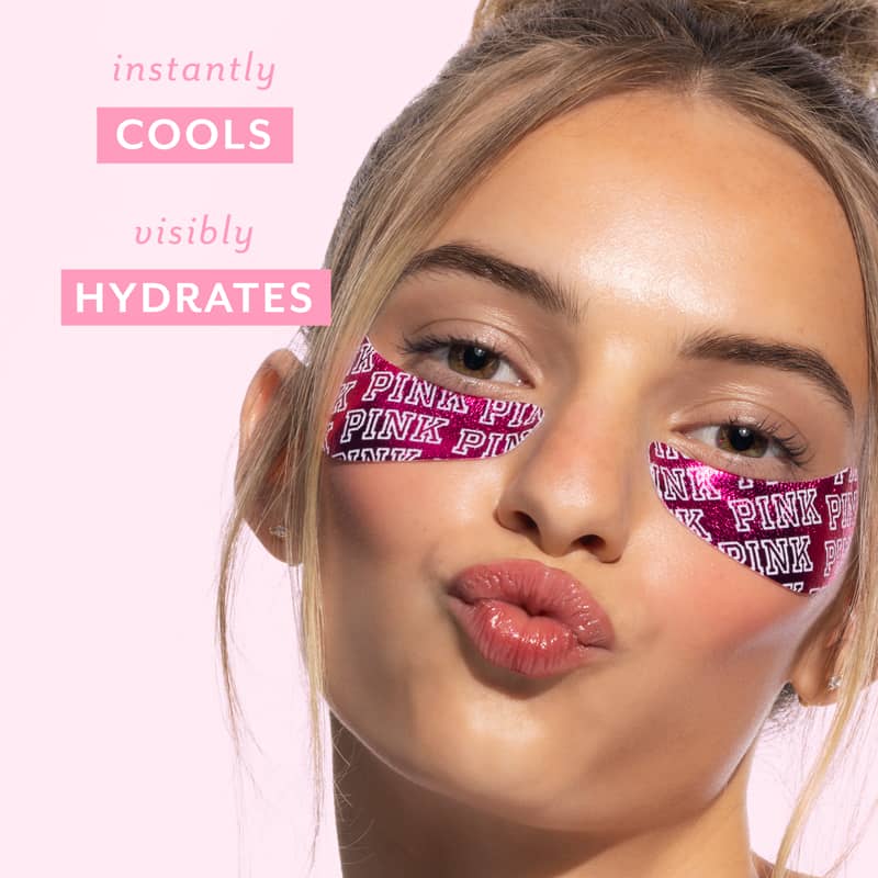 A close-up of a woman with pink Victoria's Secret PINK eye tape applied to her under-eye areas, set against a solid light pink background with promotional text that reads 'instantly COOLS visibly HYDRATES'.