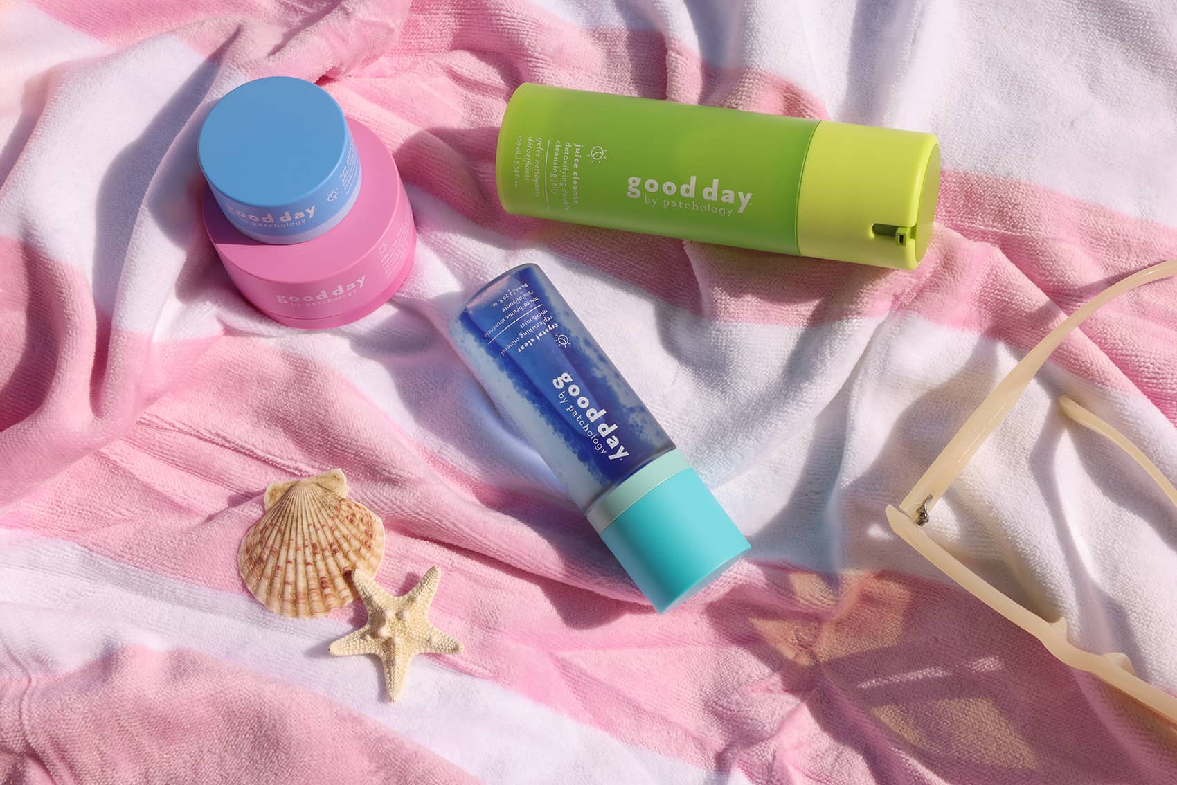 Three Good Day sunscreen products arranged on a pink and white striped beach towel with seashells and sunglasses as props: a lime green tube, a blue-and-pink jar, and a blue tube with turquoise cap.