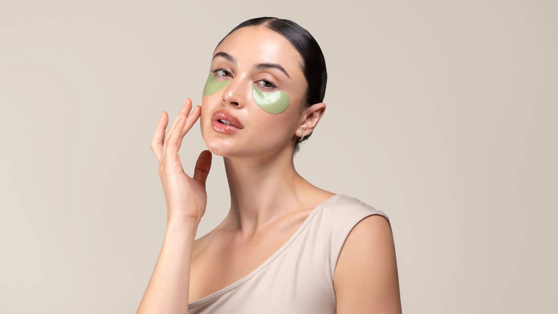 Woman applying green cool crush under eye patches on a beige background