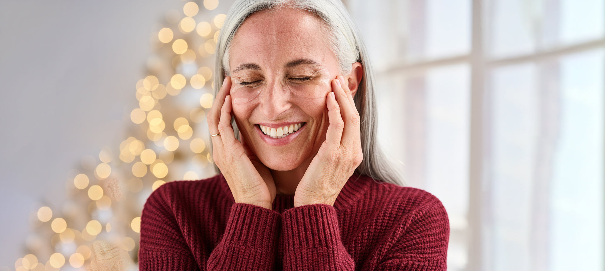 Woman in a red sweater smiling with hands on her face, Christmas tree in the background
