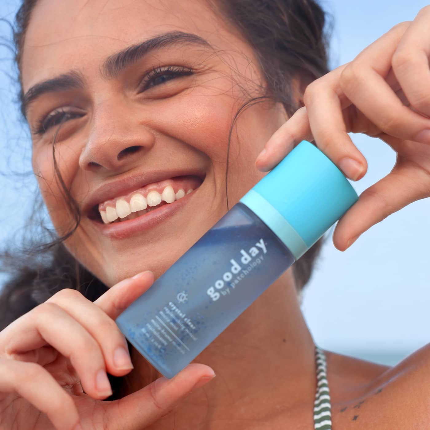 A smiling woman outdoors holds a blue and light blue bottle crystal clear against a pale sky background.
