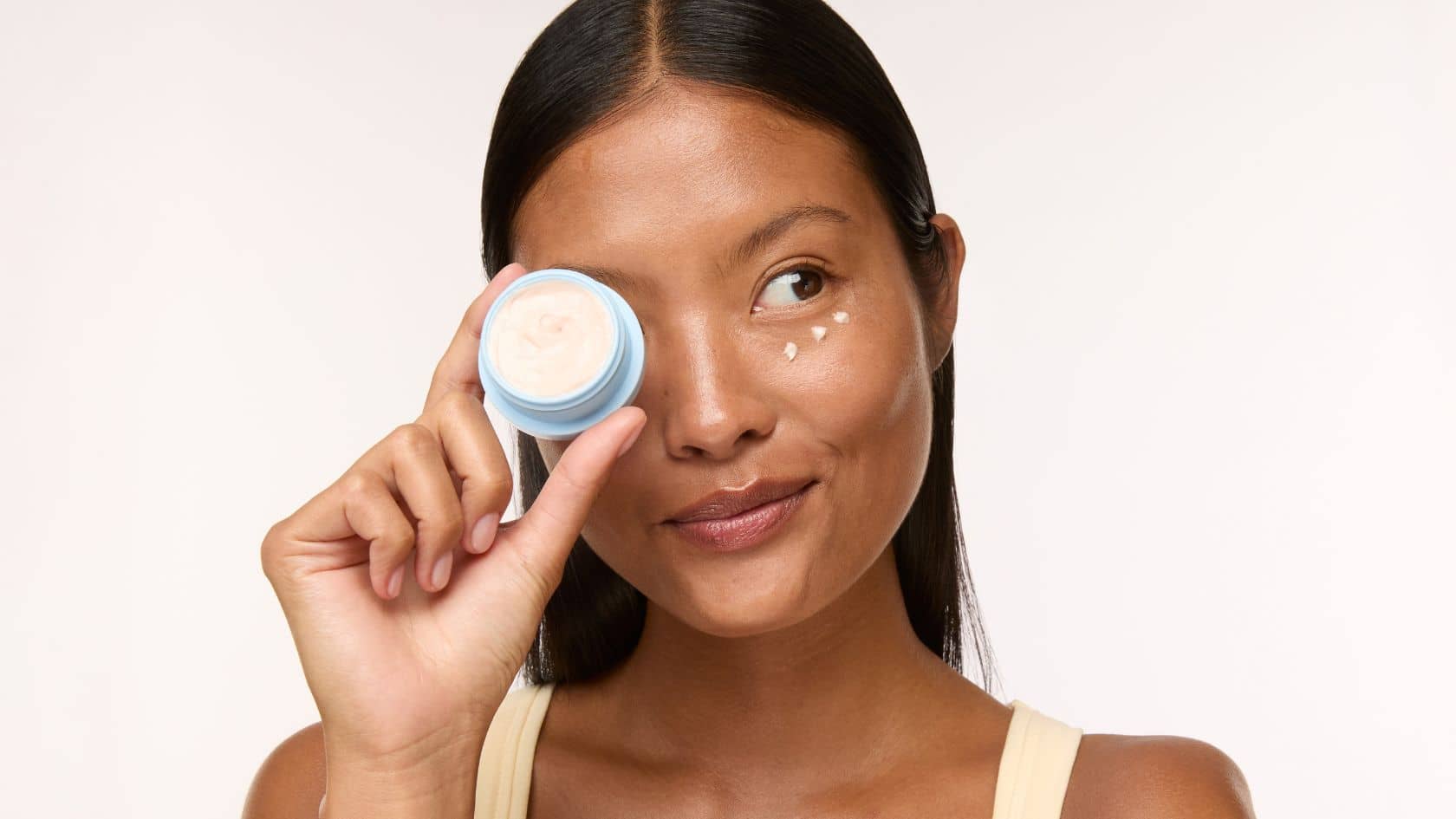 A woman holding a small round cosmetic jar near her eye, with a few drops of cream dotted on her cheek against a plain light background.