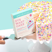 Patchology Eye Want Cake eye patches box beside a sprinkle-covered cake slice on a stand, highlighting a playful, dessert-inspired brightening and hydrating under-eye treatment

