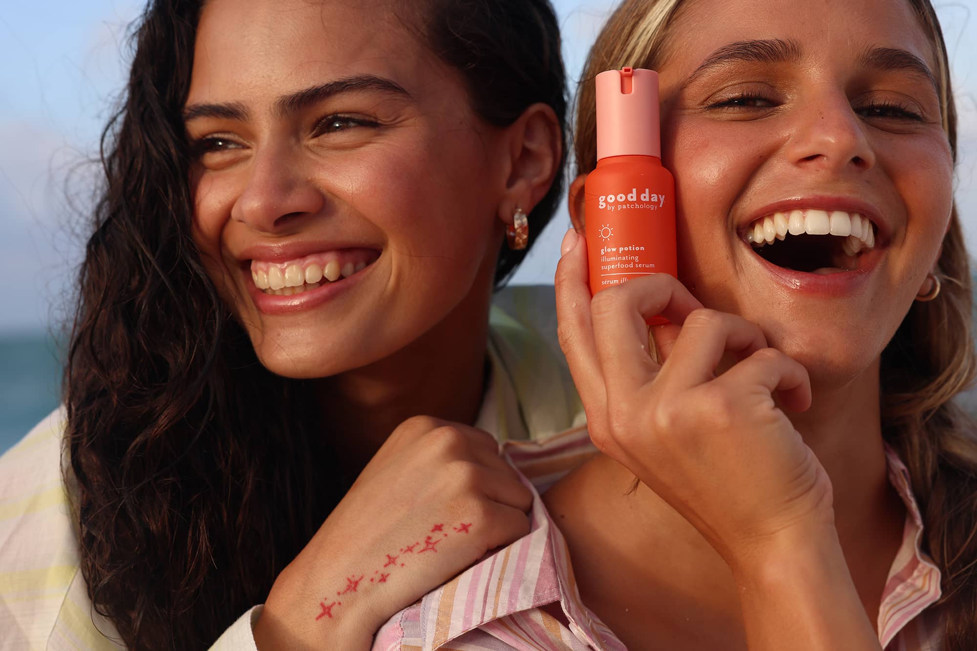 Two smiling women outdoors near the beach hold a small orange glow potion bottle labeled 'good day'.