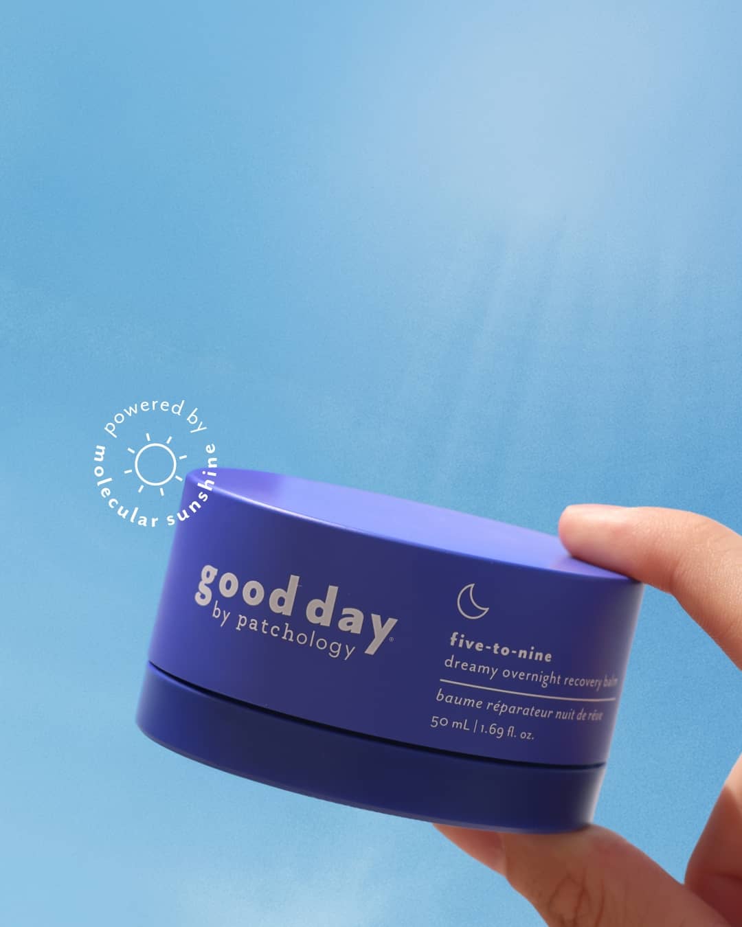 A hand holding a blue jar of 'good day' five-to-nine dreamy overnight recovery balm against a blue sky-like background.