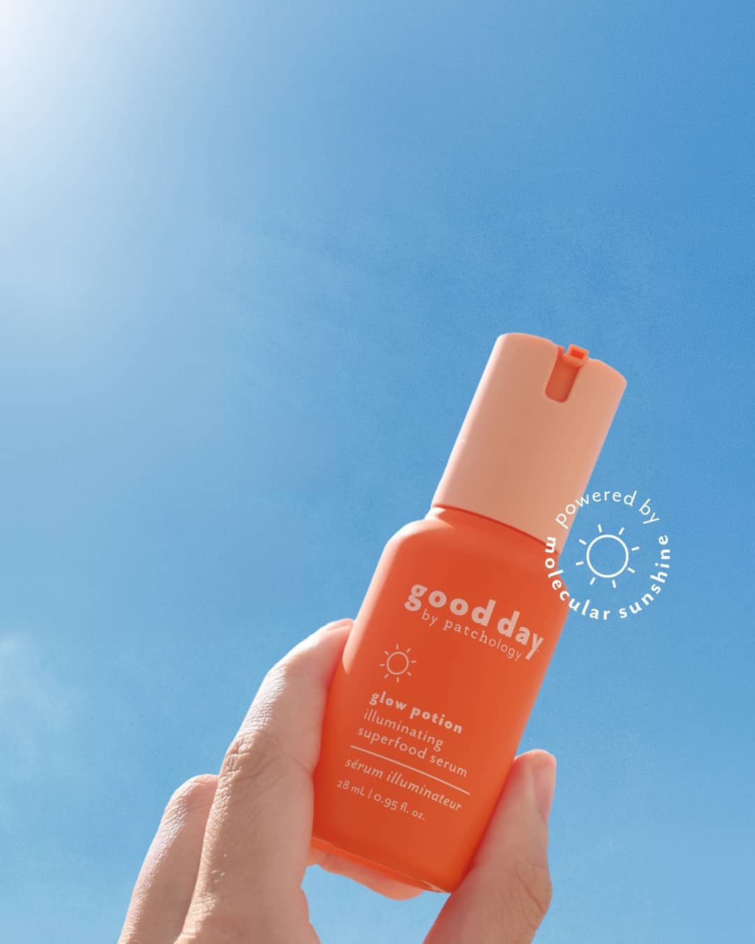 A hand holding an orange bottle of Good Day glow potion facial serum against a bright blue sky background.
