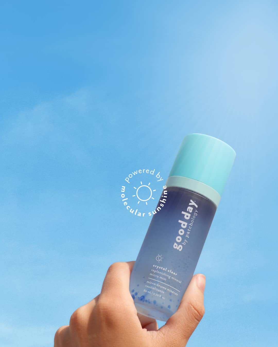 A hand holding a bottle of Good day Skincare Crystal Cleanser Facial mist against a bright blue sky background. The bottle is dark blue with a light blue cap, and a circular watermark reading 'powered by molecular sunshine' is visible on the left side.