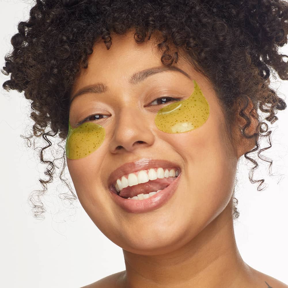 Smiling woman with curly hair wearing green, speckled hydrogel eye patches for a refreshing and depuffing skincare routine.