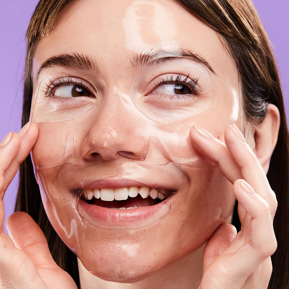 Smiling woman smoothing a clear sheet mask over her face against a purple background.
