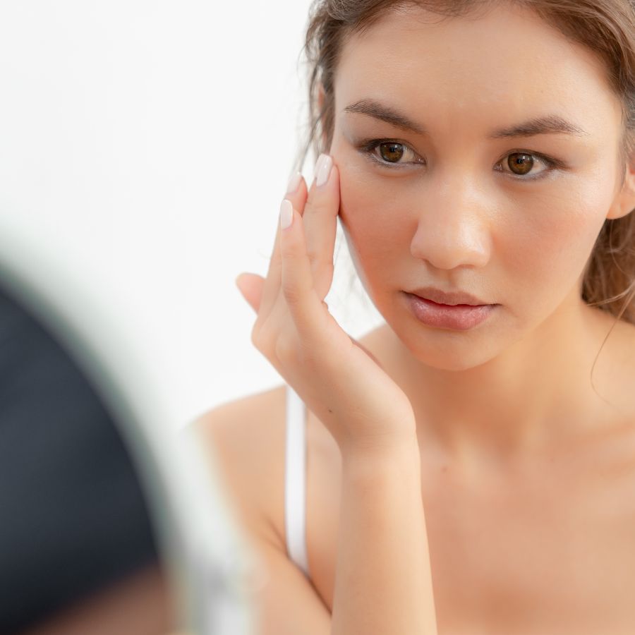 Woman examining her skin in a mirror with a neutral background