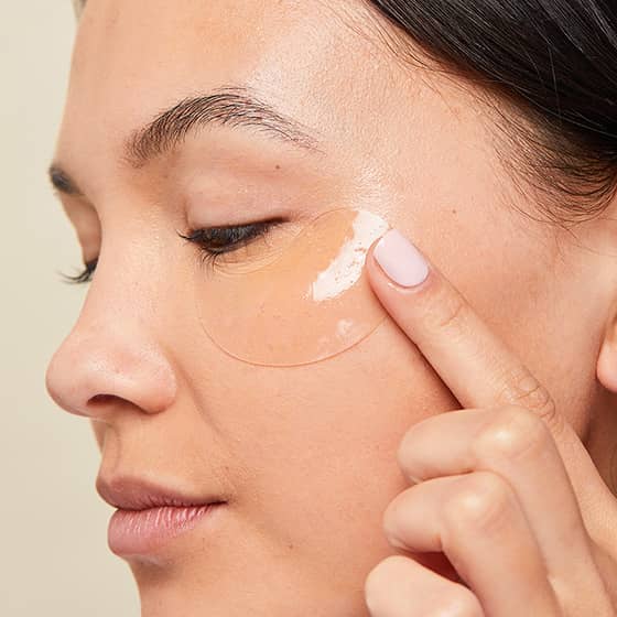 Woman pressing a clear under-eye gel patch onto her cheek during a skincare routine.

