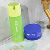 Two Good Day skincare products on a marble bathroom counter: a bright green juice cleanser bottle and a royal blue cream jar, with gold rings on a mint tray nearby.