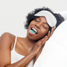A woman with dark hair laying on a pillow, wearing a silk sleep mask on her head with her eyes closed and a Little Helpers® Supplement Strips: Sleep on her tongue
