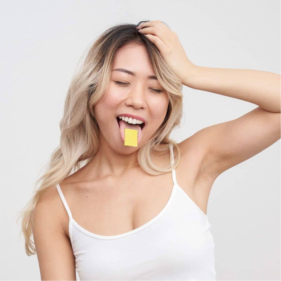 Model with long brown-blonde hair has her arm up and a Little Helpers® Supplement Strips: Energy on her tongue that's sticking out
